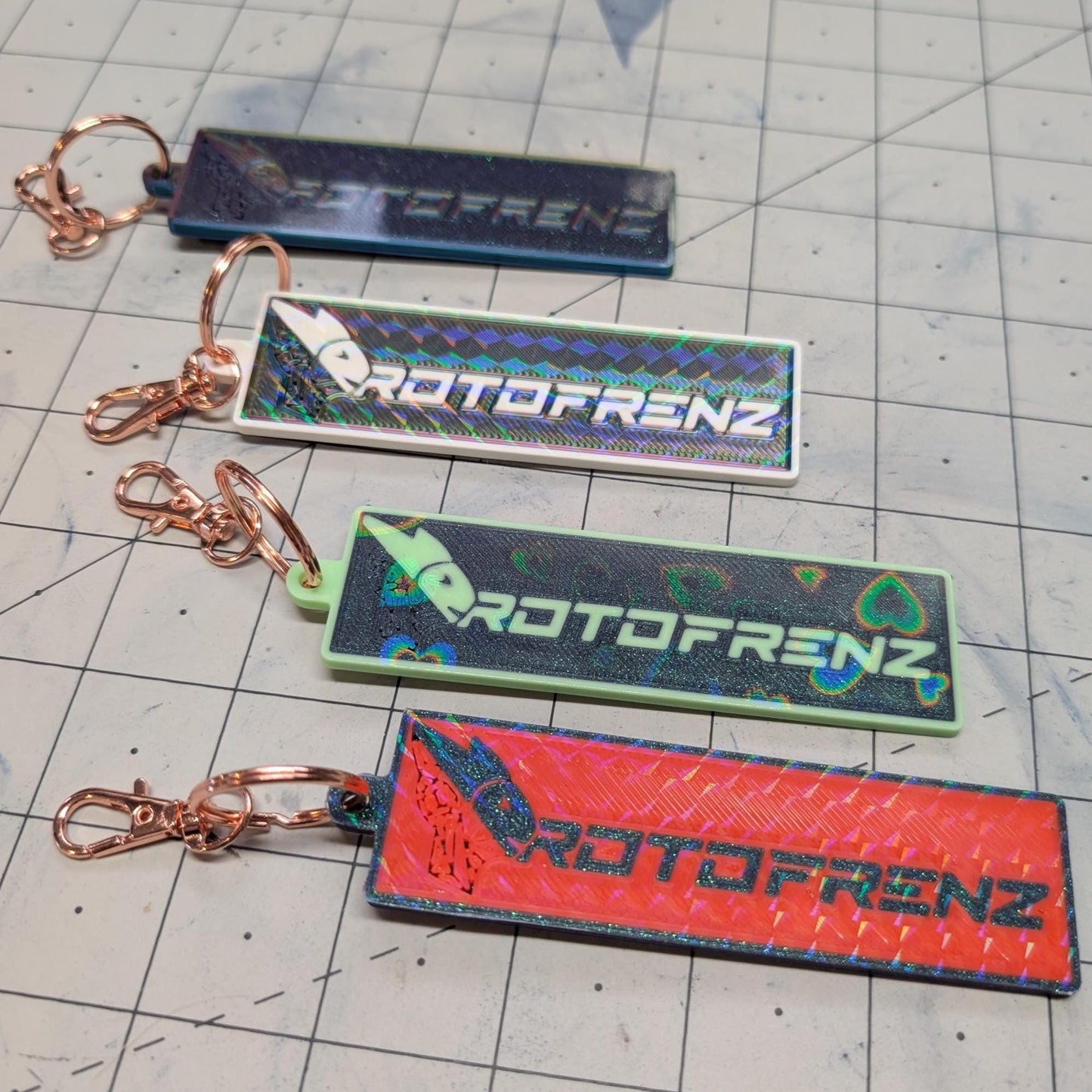 May include: Four keychains with the text 'PROTOFRENZ' printed on them. The keychains are all different colors: blue, white, green, and red. The text is in a rainbow holographic font.