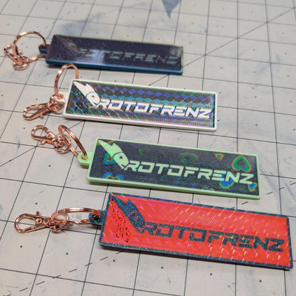 May include: Four keychains with the text &#39;PROTOFRENZ&#39; printed on them. The keychains are all different colors: blue, white, green, and red. The text is in a rainbow holographic font.
